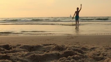 Sörf tahtası olan küçük çocuk gün batımında sahilde sörf öğreniyor.