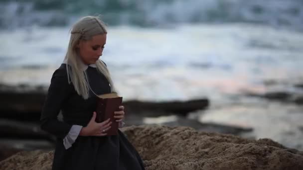 Piękne blond kobieta w staromodny sukienka z książką w ręku jest Oglądanie zachodu słońca nad morzem. — Wideo stockowe