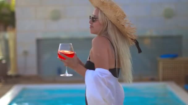 Mädchen in schwarzem Badeanzug und Strohhut tanzt mit einem Cocktail in der Hand am Pool — Stockvideo