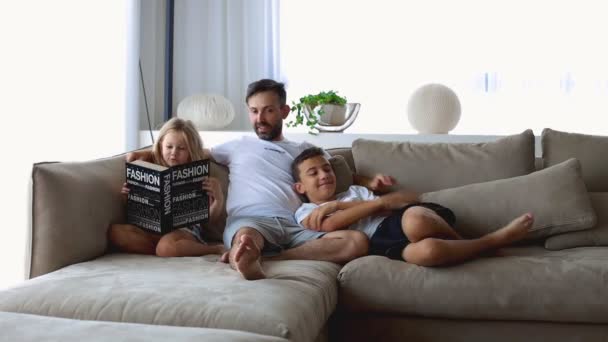 2人の子供が家のリビングルームで一緒に本を読んでいる幸せな家族 — ストック動画