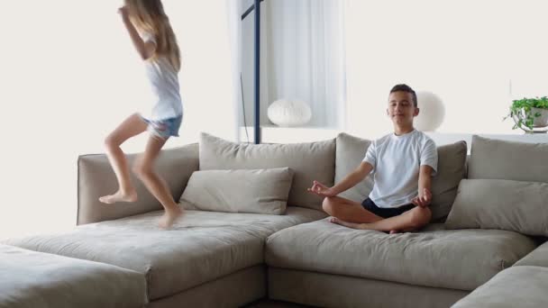 Пока активная сумасшедшая сестра прыгает рядом с братом он сидит в позе лотоса практика самоконтроля чувствовать спокойствие — стоковое видео