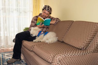 Yetişkin bir adam kanepede evcil hayvanının yanında kitap okuyor.