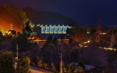 Aracın ışıkları ve arka planında hidroelektrik barajı olan gece vakti otoyolu.