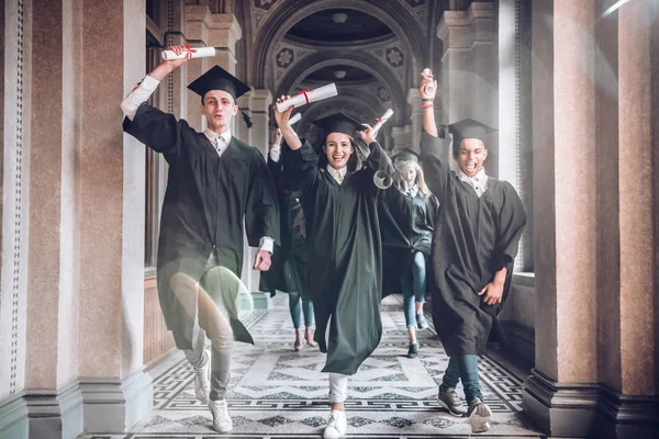 Celebrating their achievements together.University was the best years of their lives!Group of smiling university students holding their diplomas and running after being graduated in university hall.