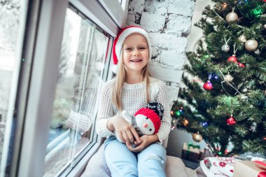 Portre sevimli mutlu küçük kız Noel kırmızı şapka takıyor. Pencere festively tatil detayları ile dekore edilmiş, komik çocuk oturur