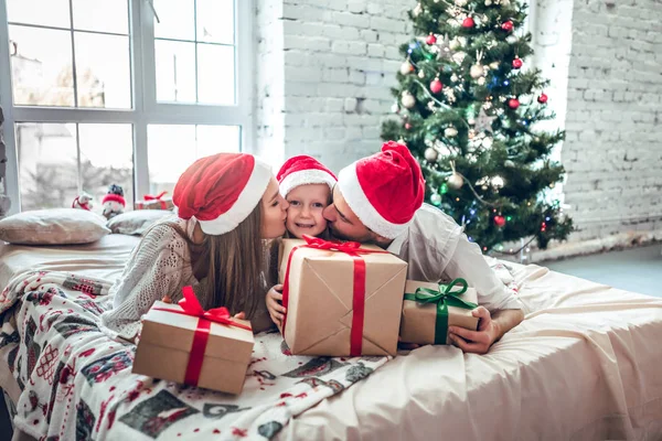 Happy parents kissing daughter and giving xmas present on bed. Christmas tree background