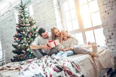 Noel. Aile. Mutluluk. Baba annesinin dizinin üstüne oturan kızı hediye veriyor. Noel ağacı yakın evde, tüm gülümsüyor musun
