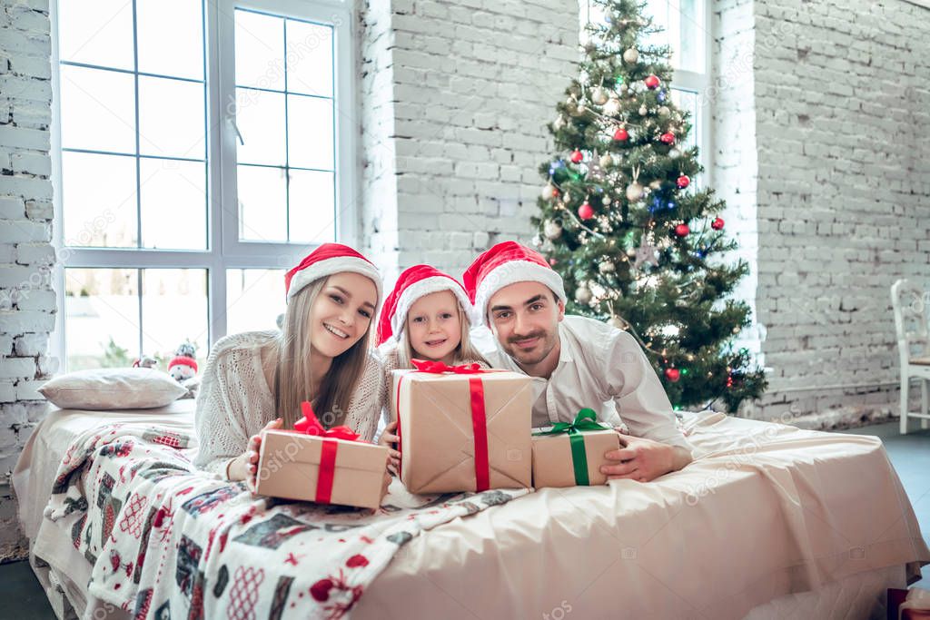 Family in Christmas Santa hats lying on bed. Mother father and baby having fun in bedroom. People relaxing at home. Winter holiday Xmas and New Year concept