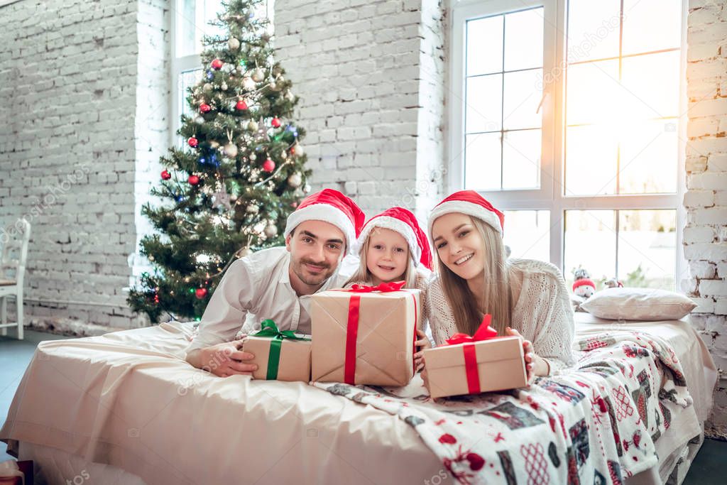 Family in Christmas Santa hats lying on bed. Mother father and baby having fun in bedroom. People relaxing at home. Winter holiday Xmas and New Year concept
