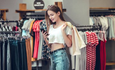 Güzel kız alışveriş torbaları tutarak, bir akıllı telefon kullanarak ve çarşıda alışveriş yaparken gülümseyen.