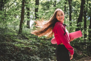 Bir spor mat bir orman yolu üzerinde tutan bir kukuleta ile sportif bir kazak içinde çırpınan saçlı genç sporcu kadın resim kırpılmış.