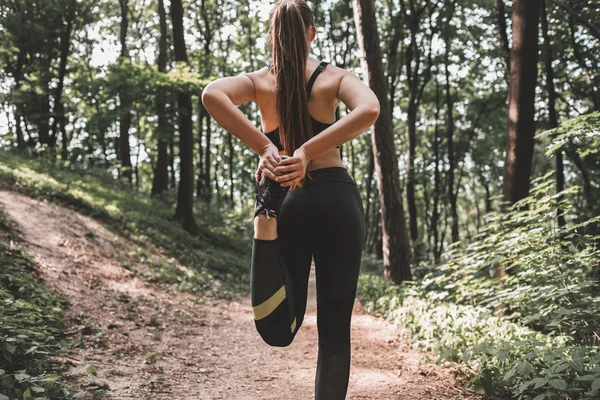 Cropped image of young woman athlete making warm-up before training in the woods. Back view of a slim fit female preparing to run on forest path. Sporty girl stretching leg before jog