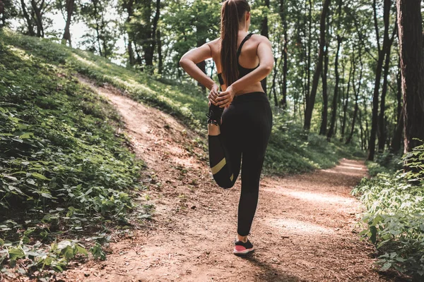 Full length image of young woman athlete making warm-up before training in the woods. Back view of a slim fit female preparing to run on forest path
