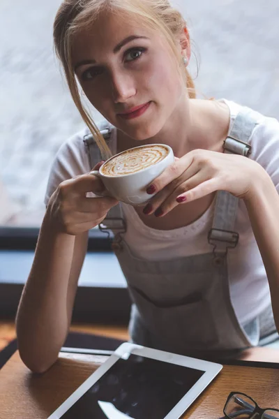 Enjoying hot coffee. Top view of an attractive young woman looking at camera and smiling while holding a cup of coffee in cafe.