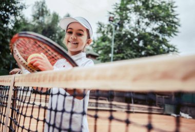 Eğitim genç çocuk tenis kulübü için. Kameranın yakınındaki tenis net bakarak küçük kız atlet portresi. Sahasında gülümseyen sportif tenis oyuncusu. Çocuklar için yaz etkinlikleri.