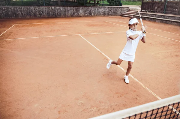 Child learning to play tennis in the sport club. Full length shot of a little girl tennis player on clay court. Sporty child girl hits the ball with a racket. Tennis training for young kid.