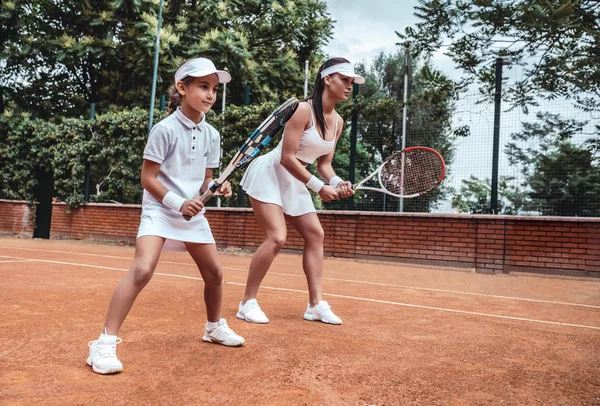 Tennis training. Cheerful mother in sports clothing teaching his daughter to play tennis while both standing on tennis court. side view