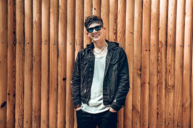 Açık havada duran ve duvara yaslanmış şık yakışıklı genç adam portresi. Seyir ve kameraya gülümseyen ceplerinde denim ceket, güneş gözlüğü, gömlek ve tutan eller giyen adam.