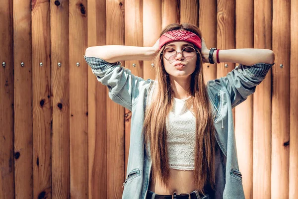 Colorful portrait of young hipster woman. Crazy girl in red bandage and rock sunglasses holding her head. Hipster woman near wooden background.