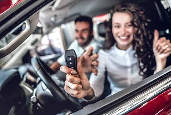 Close up of attractive woman showing car key through the window. Nice young couple sitting inside the new car at the dealership. Focus on key