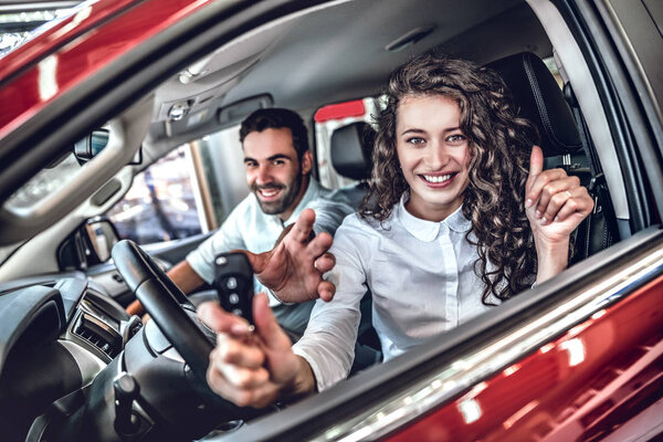 Close up of a happy young couple smiling showing car keys and thumbs up in a new car at the dealership. selective focus