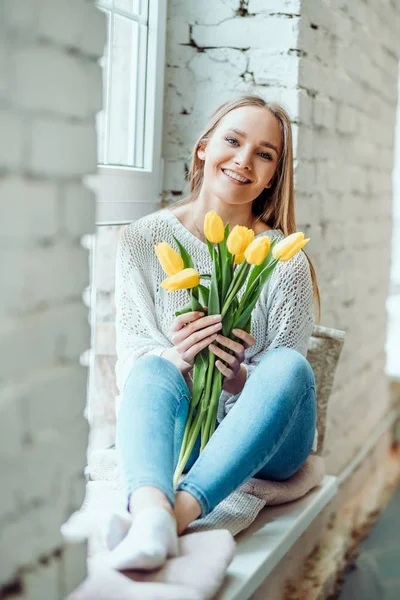 Schoonheid en tederheid concept. Portret van mooie vrouw met boeket van gele tulpen zittend op de vensterbank en camera te kijken. — Stockfoto