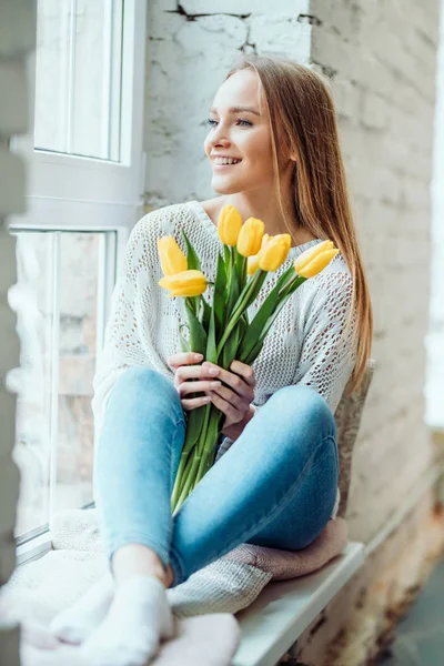 Beauty and tenderness concept.Portrait of beautiful woman with bouquet of yellow tulips sitting on the window sill and looking side.