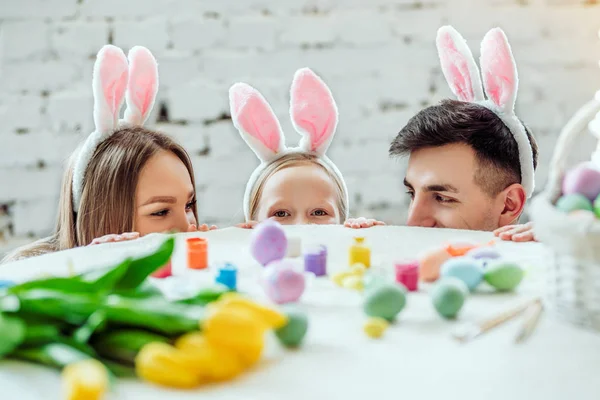 Lasst uns zusammen spielen. Schöne Familie bereitet sich gemeinsam auf Ostern vor. glückliche Mutter, Tochter und Vater verstecken sich am Tisch und schauen einander an. — Stockfoto