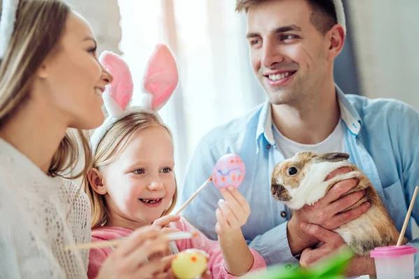 Glückliche Familie bereitet sich auf das Osterfest vor. Mutter und Tochter bemalen Eier, Papa hält ein dekoratives Kaninchen zu Hause. — Stockfoto