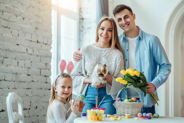 Happy easter!Dad holds a bouquet of tulips, mom holds a home decorative rabbit, daughter paints eggs and looking at camera.Celebration concept. — Stock Photo, Image