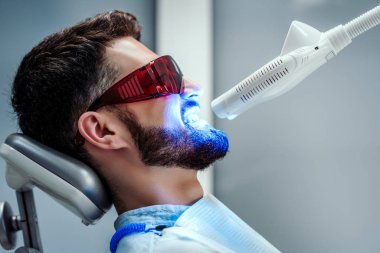 Modern klinikte diş sandalyesinde oturan genç adamın dişlerini beyazlatma tanınmaz diş hekimi. Yan görünüm.