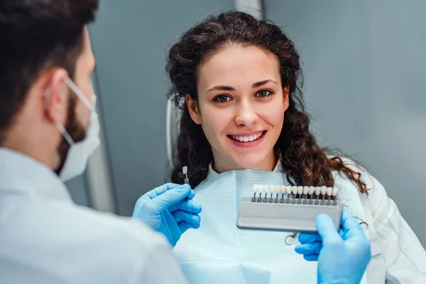 Choix de la couleur des dents avec une échelle spéciale. Dentiste choisir une nuance d'émail dentaire pour la jeune jolie fille de la patiente. Blanchiment, prothèses, dentisterie orthopédique . — Photo