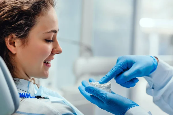 Il dentista maschile spiega la sua paziente femminile sulla cura dei denti, mostrando la sua muffa dentale. Un dentista professionista che lavora nel suo studio. Donna dal dentista per un controllo orale. Comunicazione, trattamento . — Foto Stock