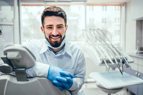 Vue de face du dentiste professionnel masculin en manteau de médecin blanc et gants de protection assis dans la chaise et l'équipement dentaire, regardant la caméra et souriant. Homme barbu posant pendant le processus de travail . — Photo