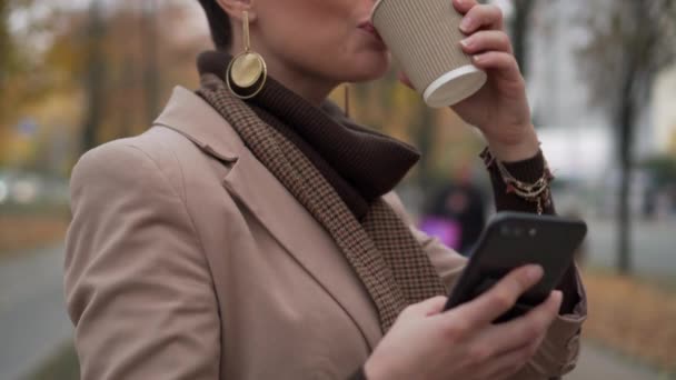 Ung kvinna dricker kaffe och använder smartphone på gatan — Stockvideo