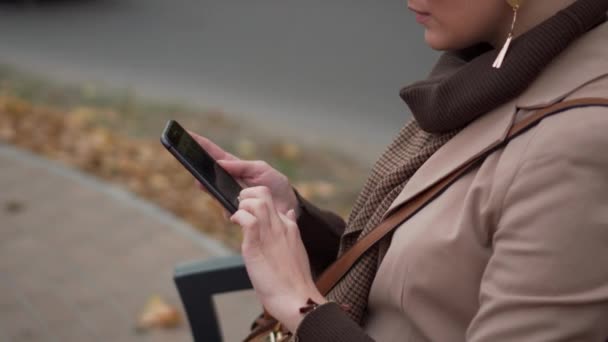 Mujer joven utiliza el teléfono inteligente mientras está sentado en el banco de la ciudad — Vídeos de Stock