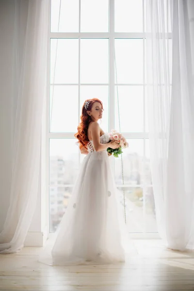 Novia pelirroja en vestido de novia blanco se encuentra cerca de una gran ventana — Foto de Stock