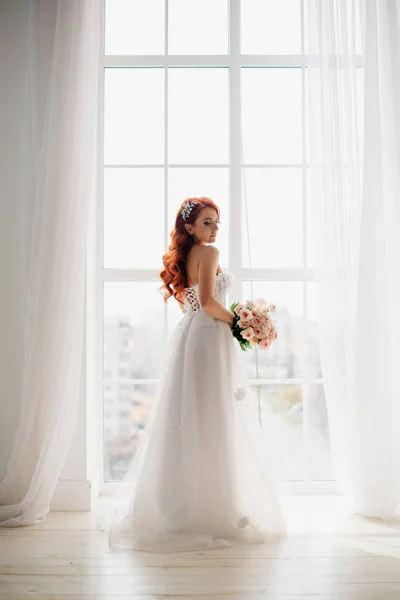 Novia pelirroja en vestido de novia blanco se encuentra cerca de una gran ventana — Foto de Stock