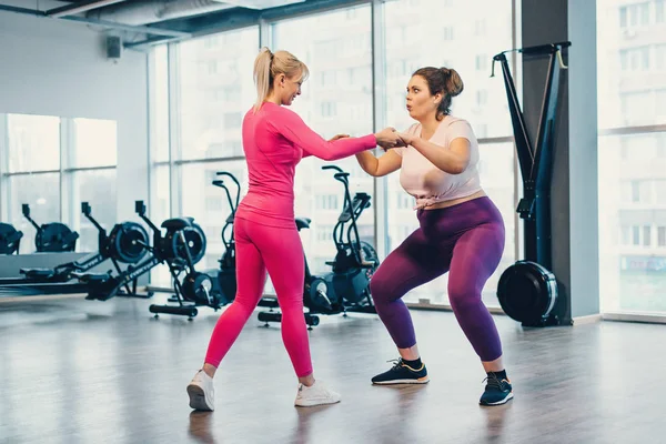 Fat woman squats in gym under supervision of trainer