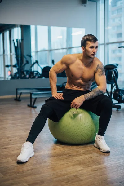 Çıplak göğüslü genç atletik adam jimnastik topunda oturuyor.