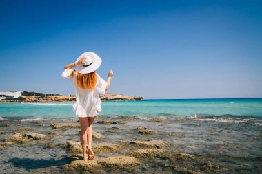 Beyaz elbiseli kızıl saçlı kadın Akdeniz kıyılarında kameraya sırtını dönüyor