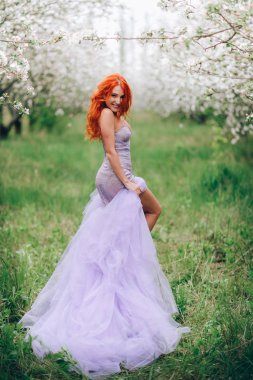 Lüks elbise güzel kızıl saçlı kadın yemyeşil bahar bahçesinde duruyor