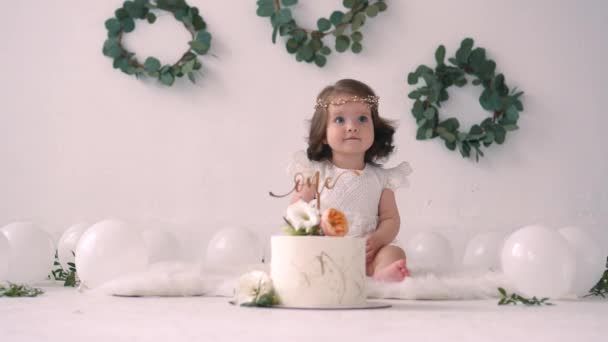 Bambina in abito bianco seduta sul pavimento vicino torta di compleanno il suo primo compleanno — Video Stock