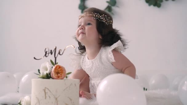 Glückliches Baby im weißen Kleid sitzt neben festlicher Torte auf ihrer ersten Geburtstagsparty, umgeben von Luftballons — Stockvideo