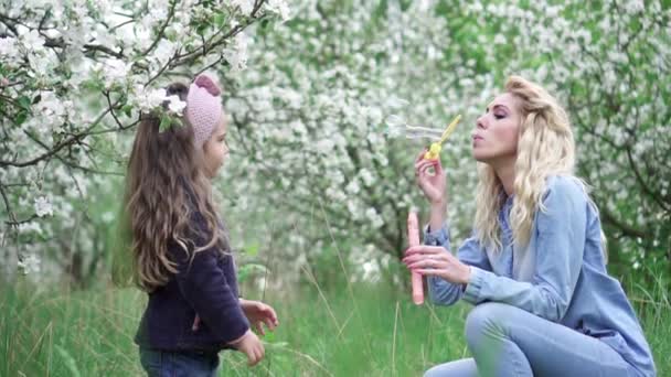 Gelukkige moeder en baby dochter spelen met zeepbellen in bloeiende tuin, Slow Motion — Stockvideo