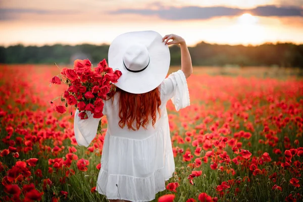Mulher ruiva de chapéu branco fica no campo florido de papoilas vermelhas com as costas para a câmera — Fotografia de Stock