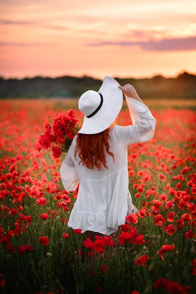 Mulher ruiva de chapéu branco fica no campo florido de papoilas vermelhas com as costas para a câmera — Fotografia de Stock