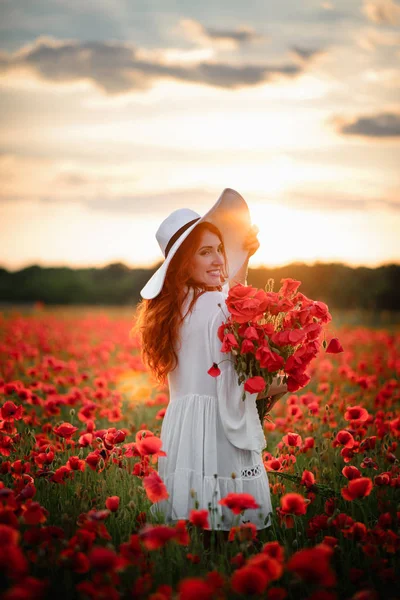 Jovem bela fêmea com buquê de papoilas vermelhas fica no campo florido ao pôr do sol — Fotografia de Stock