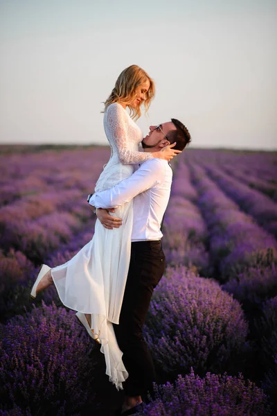 Joyeux marié tenant sa mariée dans ses bras dans un champ de lavande en fleurs au coucher du soleil — Photo