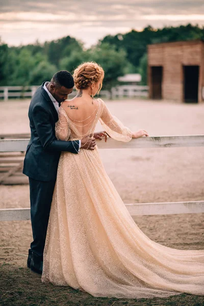 Two lovers man and woman stand embracing on a ranch at sunset, man kisses woman neck